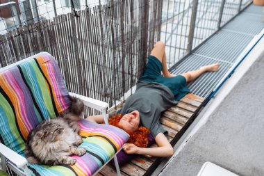 Kızıl saçlı, kıvırcık saçlı, balkonda tahta döşeme tahtasına uzanmış hiçbir şey yapmadan eğlenen genç bir kadın. Yakınlarda evcil bir kedi var, evcil hayvanla vakit geçiriyor.