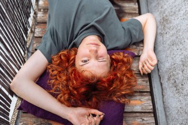 Portre kızıl saçlı kıvırcık kadın balkonda ya da terasta rahatlıyor. Kızıl saçlı, kıvırcık saçlı, balkonda tahta döşeme tahtasında uzanmış hiçbir şey yapmadan eğlenen genç bir kadın.