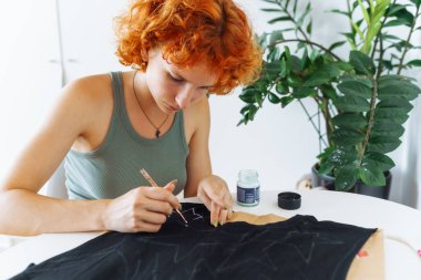 Genç bir kadın resimlerini boyalı kıyafetlere çiziyor. El yapımı el işi. Bir sanatçı, giysi üzerindeki çizgileri izlemek için fırça kullanır.