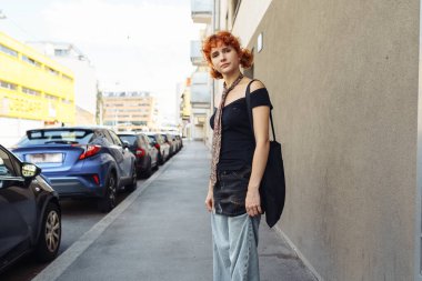 Gen z estetik kıyafeti havalı, hareketli ya da modaya uygun görünüm için. Tasarımcı markası. Kızıl saçlı genç model, olağanüstü kıyafetler, kravat, serbest stil giysiler, şehir manzarasına karşı.