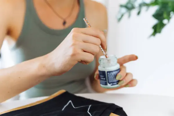 stock image young woman draws picture on clothes with paints, handmade craft work. An artist uses brush to trace lines design on clothing. close-up