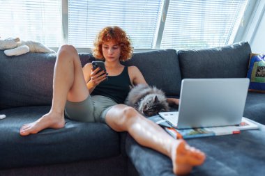 Kanepede oturan kızıl saçlı genç bir kadının portresi. Yanında uyuyan gri bir kedi. Evcil hayvanınla boş zaman geçir.