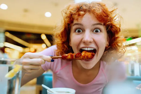 Kızıl saçlı genç kızın portresi zevkle tavuk şiş kebap yiyor. Fast food restoranında lezzetli yemekler.