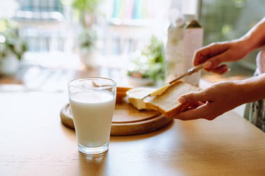 Sağlıklı kahvaltı sütlü ekmek ve tereyağı. Genç kadınların elleri kızarmış ekmeğe tereyağı sürüyor. Arka planda bir bardak taze süt var.