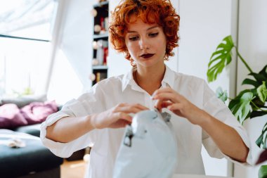 Mutlu, genç, kızıl saçlı bir kadın oturma odasında oturmuş plastik poşetleri açıyor, paket alışverişi yapıyor, internetten gülümsüyor, milenyum kızı kutusunu açıyor, internetten iyi teslimat konsepti sipariş ediyor.