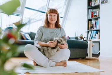 Evdeki sağlık. Orta yaşlı kadın portresi, sarışın, meditasyon yapan örgü ören halı, parke zemin, orta oda, arka plandaki kanepe, pencere. Evcil hayvanlarla yoga, kadınların kollarında kedi, birlikte.