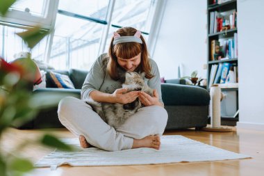 Evdeki sağlık. Orta yaşlı kadın portresi, sarışın, meditasyon yapan örgü ören halı, parke zemin, orta oda, arka plandaki kanepe, pencere. Evcil hayvanlarla yoga, kadınların kollarında kedi, birlikte.