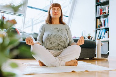 Evdeki sağlık. Orta yaşlı kadın portresi, sarışın, evde meditasyon yapan, örgü ören halı, parke döşeme, orta oda, arka plan koltuğu, pencere...