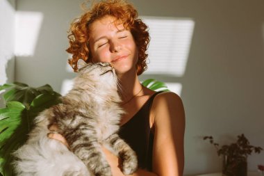 Kızıl saçlı kıvırcık kadın portresi, kollarında evcil gri tüylü kedi, güneş ışınları ve gölge, parlak güneş ışığı