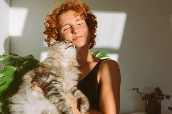 Kızıl saçlı kıvırcık kadın portresi, kollarında evcil gri tüylü kedi, güneş ışınları ve gölge, parlak güneş ışığı