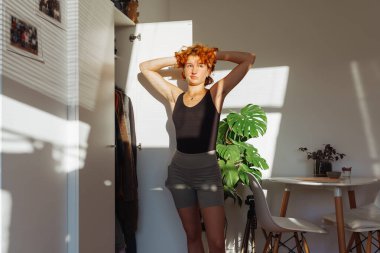 Genç kızıl saçlı çekici bir kadın, iyi organize edilmiş bir odada seçilmiş kıyafetleri değerlendiriyor, düşünceli bir ifadeyle dolabın yanında dikiliyor. Parlak ışık, duvarda güneş ışığı gölgesi, canavar bitkisi. 