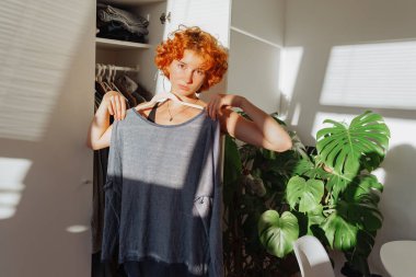 Genç kızıl saçlı çekici bir kadın, iyi organize edilmiş bir odada seçilmiş kıyafetleri değerlendiriyor, düşünceli bir ifadeyle dolabın yanında dikiliyor. Parlak ışık, duvarda güneş ışığı gölgesi, canavar bitkisi. 