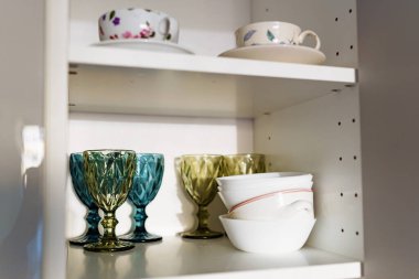 Temiz tabaklar, renkli bardaklar, mutfak dolabındaki beyaz rafta duran çatal bıçak takımı..