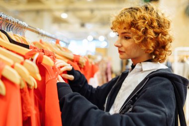 Genç ve çekici kızıl saçlı bir kadın butikte kırmızı elbiseleri seçiyor. Modern bir mağazadan kıyafet almak.