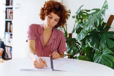 Genç, çekici, kızıl saçlı kıvırcık kadın yuvarlak masada ev kıyafetleri içinde oturuyor ve deftere notlar yazıyor.