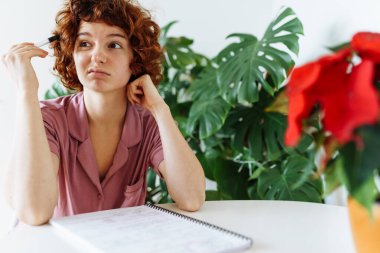 young attractive red-haired curly woman sitting in home clothes at round table, writing notes in notebook clipart