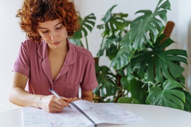 Genç, çekici, kızıl saçlı kıvırcık kadın yuvarlak masada ev kıyafetleri içinde oturuyor ve deftere notlar yazıyor.