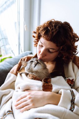 Çekici bir portre kıvırcık saçlı, sıcak bir paçavra içinde, kanepede oturuyor, gri tüylü evcil kediyi kucaklıyor.