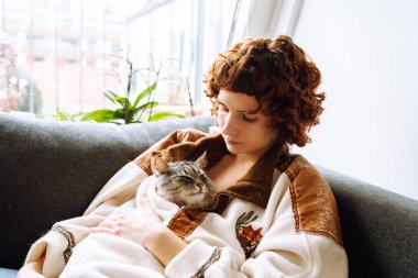 Çekici bir portre kıvırcık saçlı, sıcak bir paçavra içinde, kanepede oturuyor, gri tüylü evcil kediyi kucaklıyor.