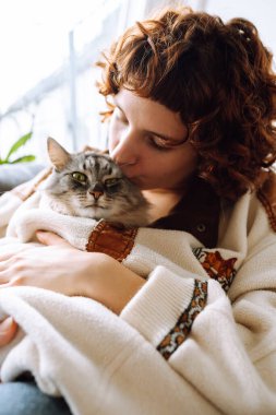 Çekici bir portre kıvırcık saçlı, sıcak bir paçavra içinde, kanepede oturuyor, gri tüylü evcil kediyi kucaklıyor.