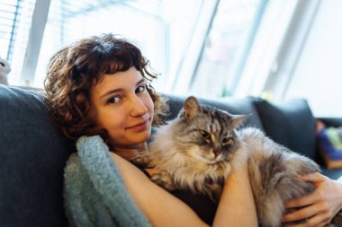 Genç, kızıl saçlı, gri tüylü kediyi kucaklayan, yumuşak koltukta oturan, battaniyeyle kaplanmış bir kadın.