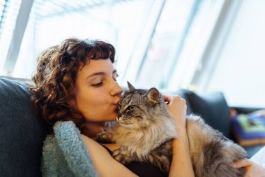 Genç, kızıl saçlı, gri tüylü kediyi kucaklayan, yumuşak koltukta oturan, battaniyeyle kaplanmış bir kadın.
