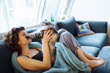 Genç, kızıl saçlı, gri tüylü kediyi kucaklayan, yumuşak koltukta oturan, battaniyeyle kaplanmış bir kadın.