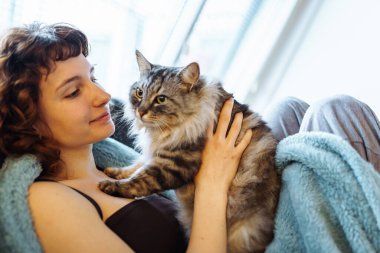 Genç, kızıl saçlı, gri tüylü kediyi kucaklayan, yumuşak koltukta oturan, battaniyeyle kaplanmış bir kadın.