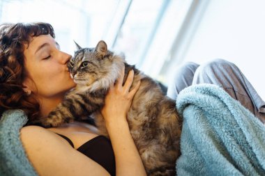 Genç, kızıl saçlı, gri tüylü kediyi kucaklayan, yumuşak koltukta oturan, battaniyeyle kaplanmış bir kadın.