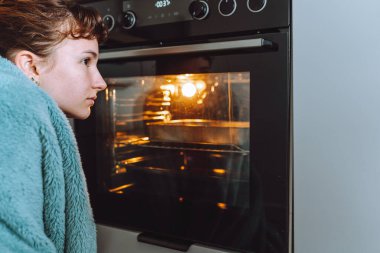 Battaniyeye sarılı genç bir kadın yemeğin hazırlanmasını ve fırında pişirilmesini izliyor.