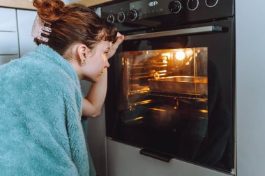 Battaniyeye sarılı genç bir kadın yemeğin hazırlanmasını ve fırında pişirilmesini izliyor.