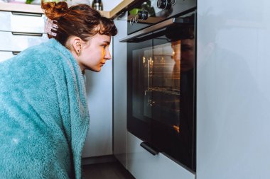Battaniyeye sarılı genç bir kadın yemeğin hazırlanmasını ve fırında pişirilmesini izliyor.