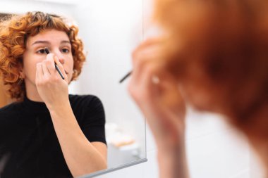 Kızıl saçlı genç kadın aynada yansıyan yüzüne makyaj yapıyor.