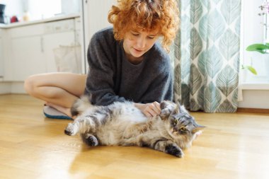 Genç, kızıl saçlı, evcil kediyi kucaklayan kadın, sıcak ahşap zeminde birlikte otururken.