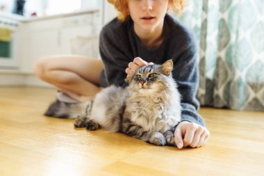 Genç, kızıl saçlı, evcil kediyi kucaklayan kadın, sıcak ahşap zeminde birlikte otururken.