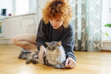 Genç, kızıl saçlı, evcil kediyi kucaklayan kadın, sıcak ahşap zeminde birlikte otururken.