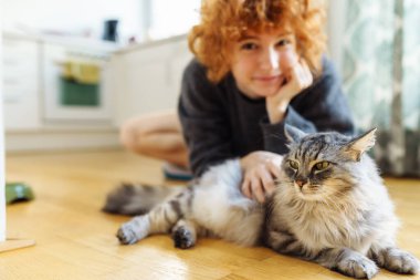 Genç, kızıl saçlı, evcil kediyi kucaklayan kadın, sıcak ahşap zeminde birlikte otururken.