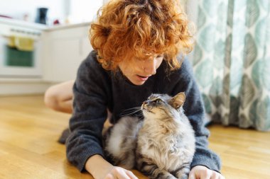 Genç, kızıl saçlı, evcil kediyi kucaklayan kadın, sıcak ahşap zeminde birlikte otururken.
