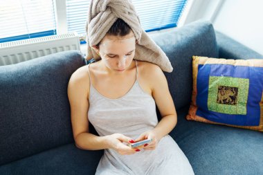 Islak saçlı genç bir kadın yumuşak rahat kanepeye oturur, pencerenin yanında, evin içinde, akıllı telefon kullanır.,