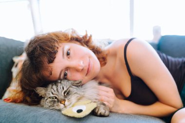 Evcil kedi ve sahibi yumuşak kanepede oturuyorlar. Kedi en sevdiği oyuncağına sarılıyor..