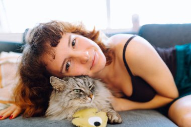 Evcil kedi ve sahibi yumuşak kanepede oturuyorlar. Kedi en sevdiği oyuncağına sarılıyor..