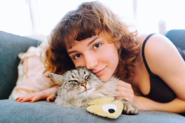 Evcil kedi ve sahibi yumuşak kanepede oturuyorlar. Kedi en sevdiği oyuncağına sarılıyor..