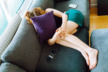 Genç bir kadın kanepeye uzanmış, başını yastıkla örtüyor, stresli.