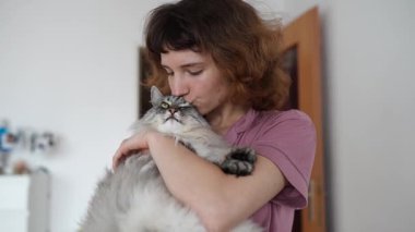 Genç kadın tüylü gri safkan kediyi kollarında tutuyor, sarılıyor, öpüyor, kedi mırlıyor, ağır çekimde mırlıyor.