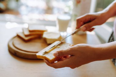 Sağlıklı kahvaltı sütlü ekmek ve tereyağı. Genç kadınların elleri kızarmış ekmeğe tereyağı sürüyor. Arka planda bir bardak taze süt var.