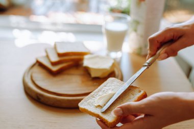 Sağlıklı kahvaltı sütlü ekmek ve tereyağı. Genç kadınların elleri kızarmış ekmeğe tereyağı sürüyor. Arka planda bir bardak taze süt var.