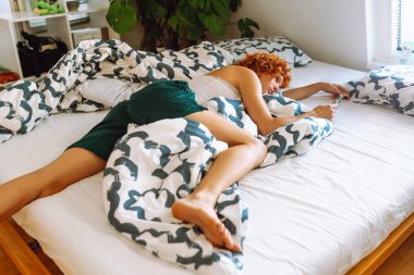 Evde yatak odasında cep telefonu kullanarak şort ve mayo giyen kızıl saçlı kıvırcık kadın portresi. Gençlerin sosyal medya bağımlılığı, telefon kullanımı.