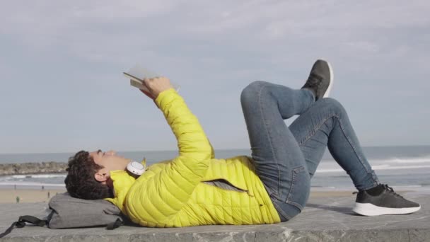 Jonge Spaanse Man Ligt Buiten Leest Een Boek Aan Zee — Stockvideo