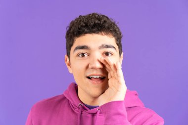 Hispanic boy whispering or telling a secret while looking at camera isolated on purple background