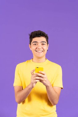Smiling teenager boy looking at camera while holding phone isolated on purple background
